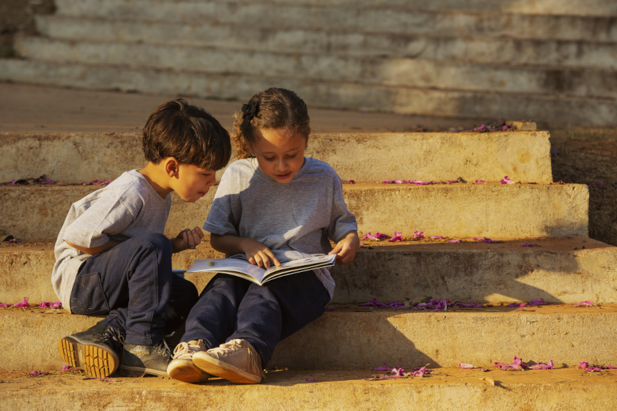 Foto de duas crianças lendo um livro sentadas em uma escada ensolarada e com algumas flores lilás ao chão